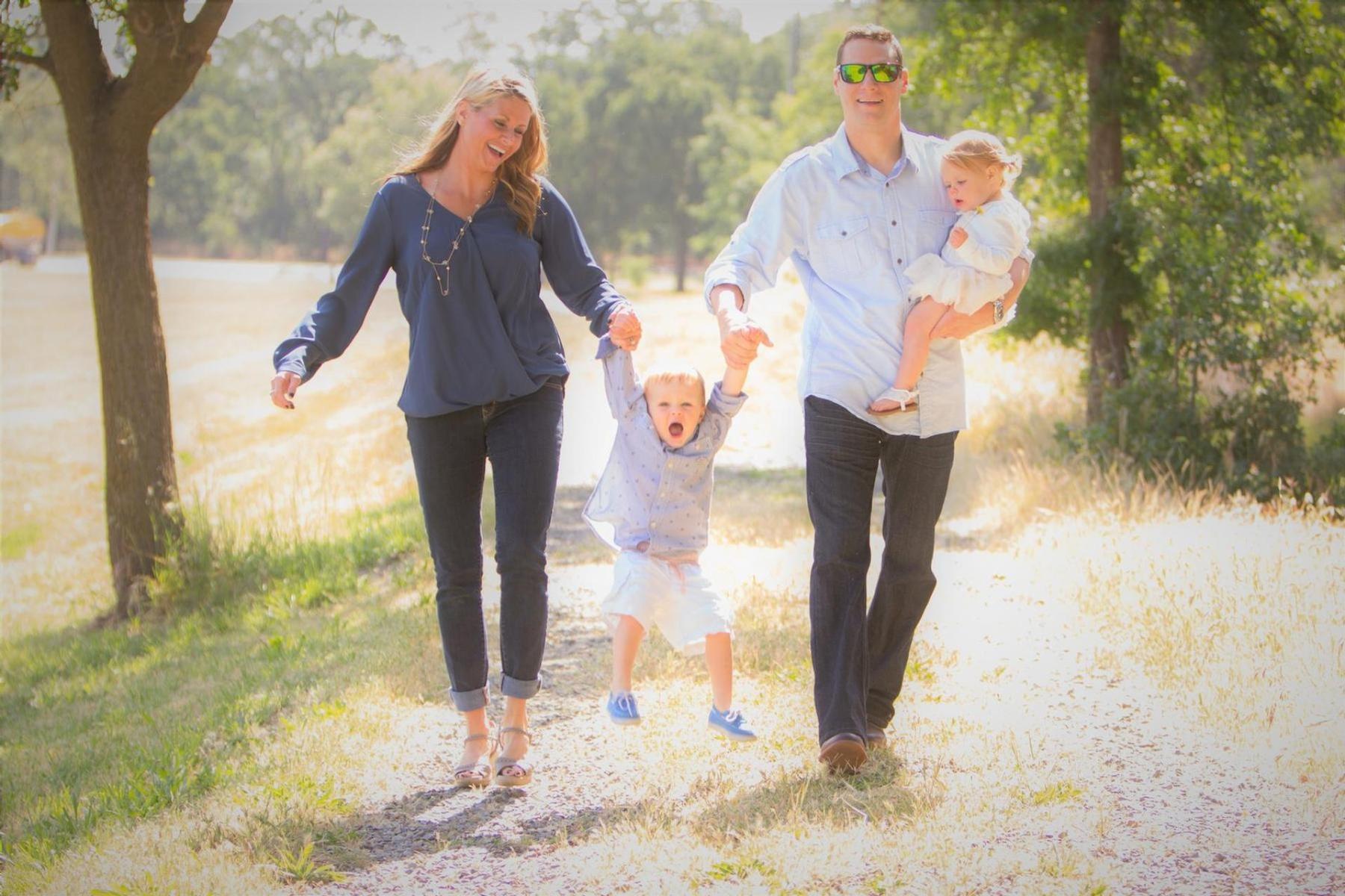 Family walking together swinging daughter on gravel road outside green trees sunshine